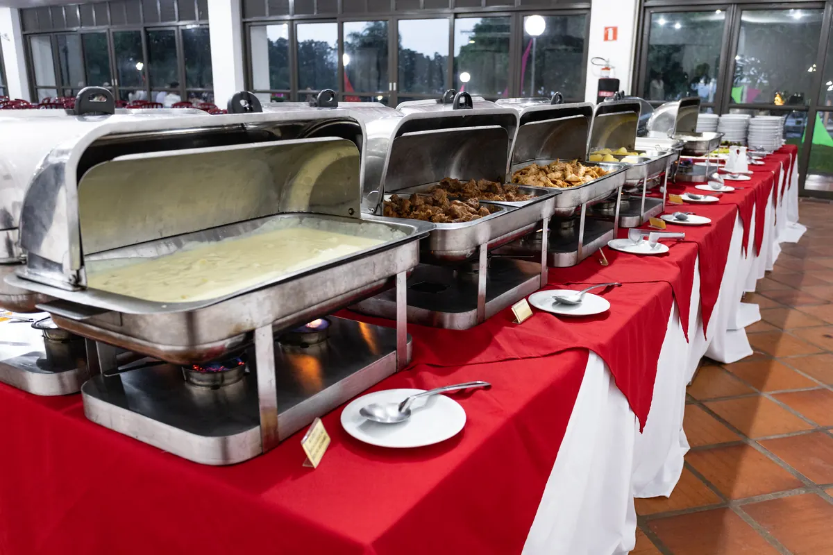 Buffet do restaurante com réchauds em linha sobre toalhas vermelhas e brancas, com janelas ao fundo.