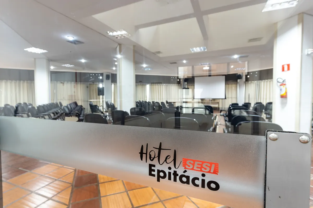 Salão de eventos com cadeiras e telão, visto através de painel de vidro com o logotipo do Hotel SESI Epitácio.