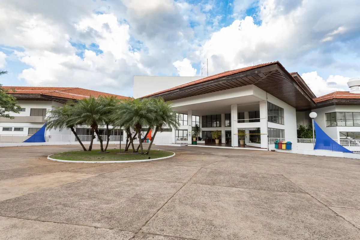 Entrada da recepção do hotel com marquise, palmeiras e céu com nuvens.