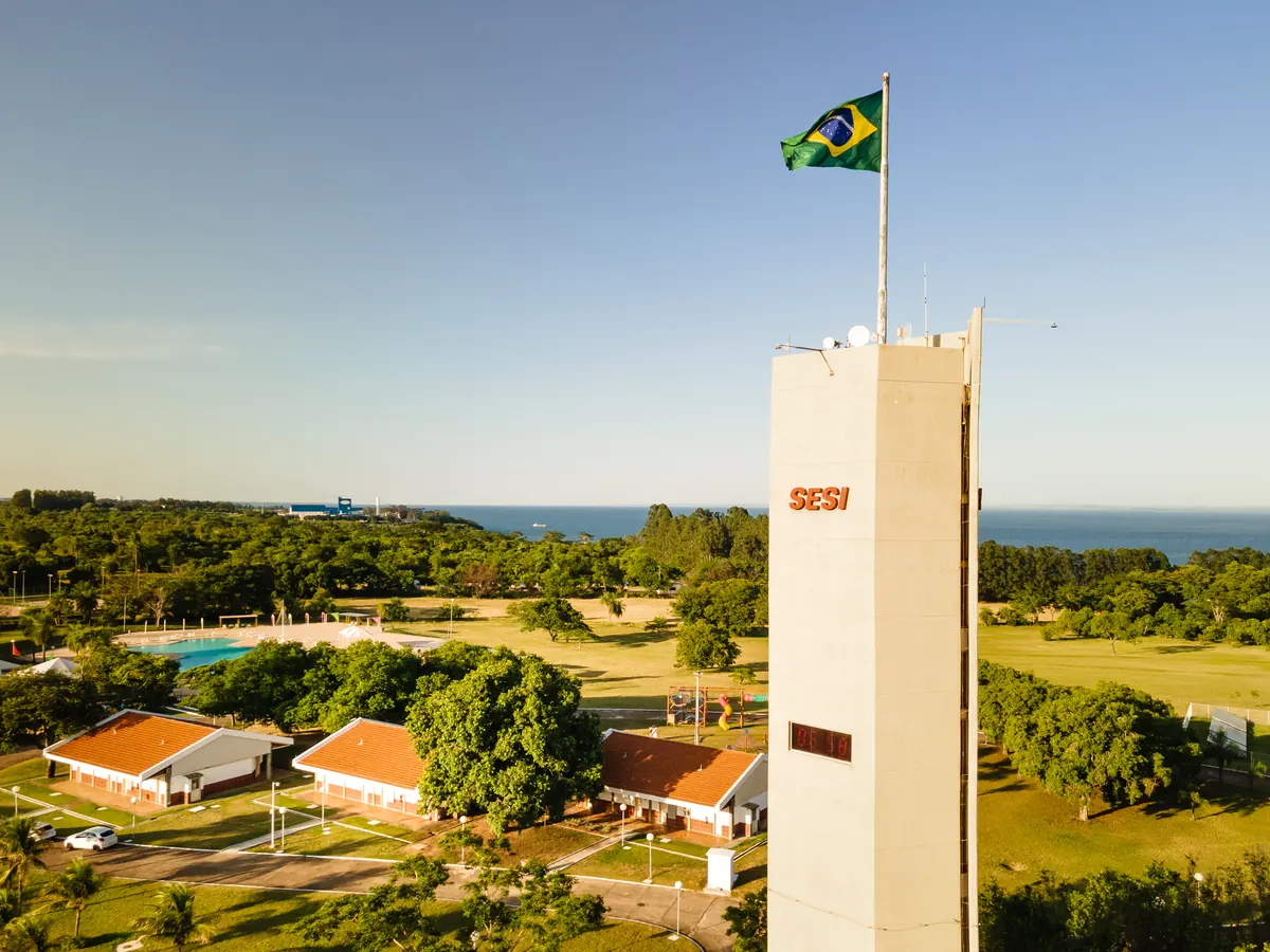 Vista aérea da torre do SESI com bandeira, bangalôs de telhado laranja entre árvores e o rio ao fundo.