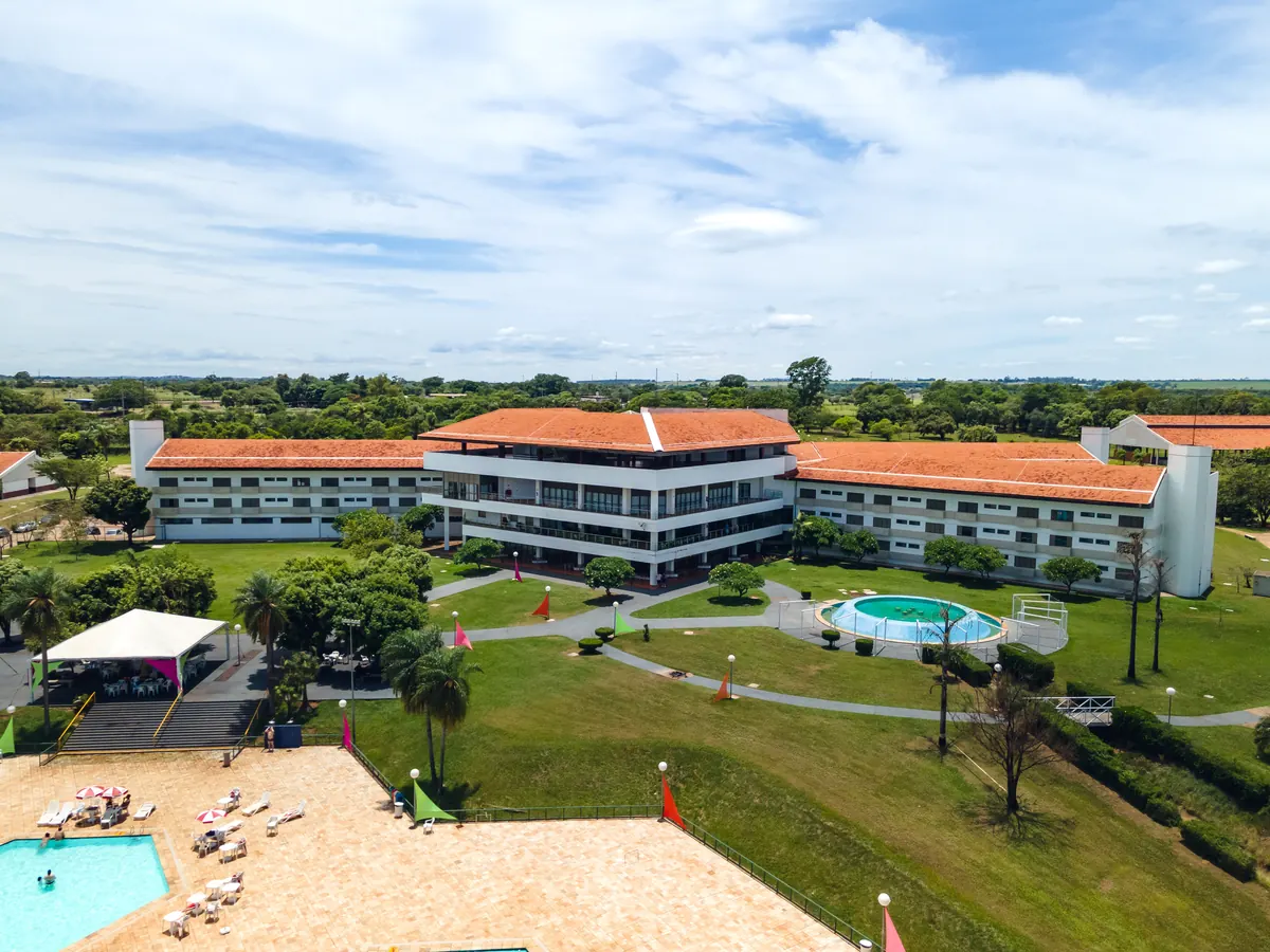 Prédio principal do hotel com jardim, fonte e canto da piscina, céu azul com nuvens