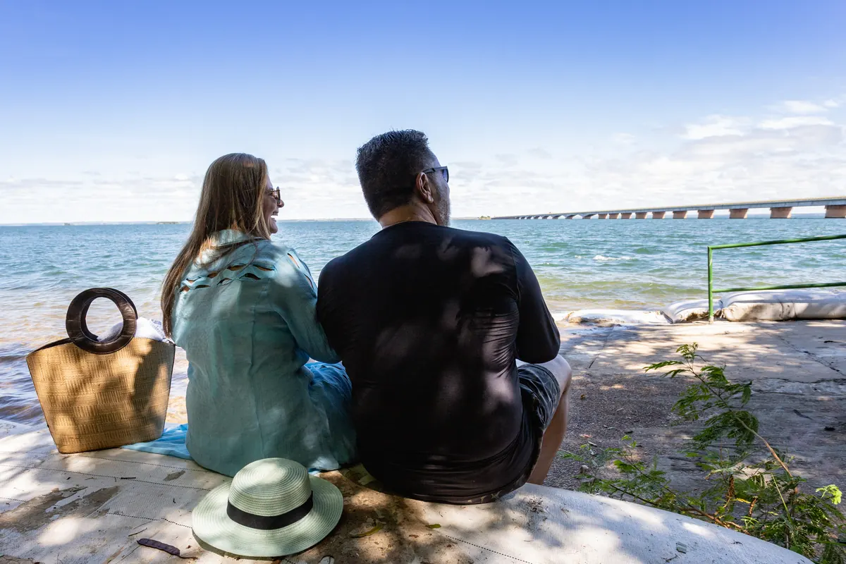 Casal contemplando o rio Paraná sentado na orla, com a ponte no horizonte