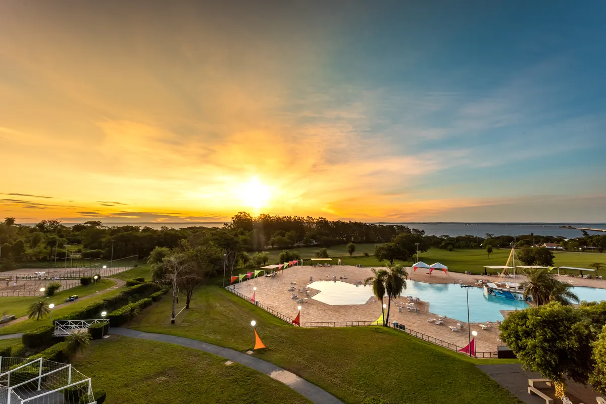 Pôr do sol com raios dourados sobre a piscina, palmeiras e o rio ao fundo