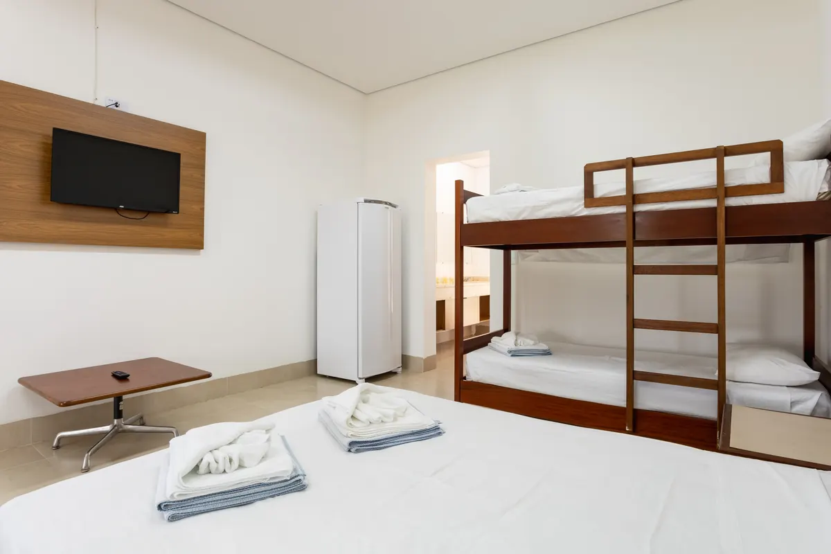 Quarto de bangalô com cama de casal, beliche, TV na parede, frigobar e toalhas dobradas sobre a cama